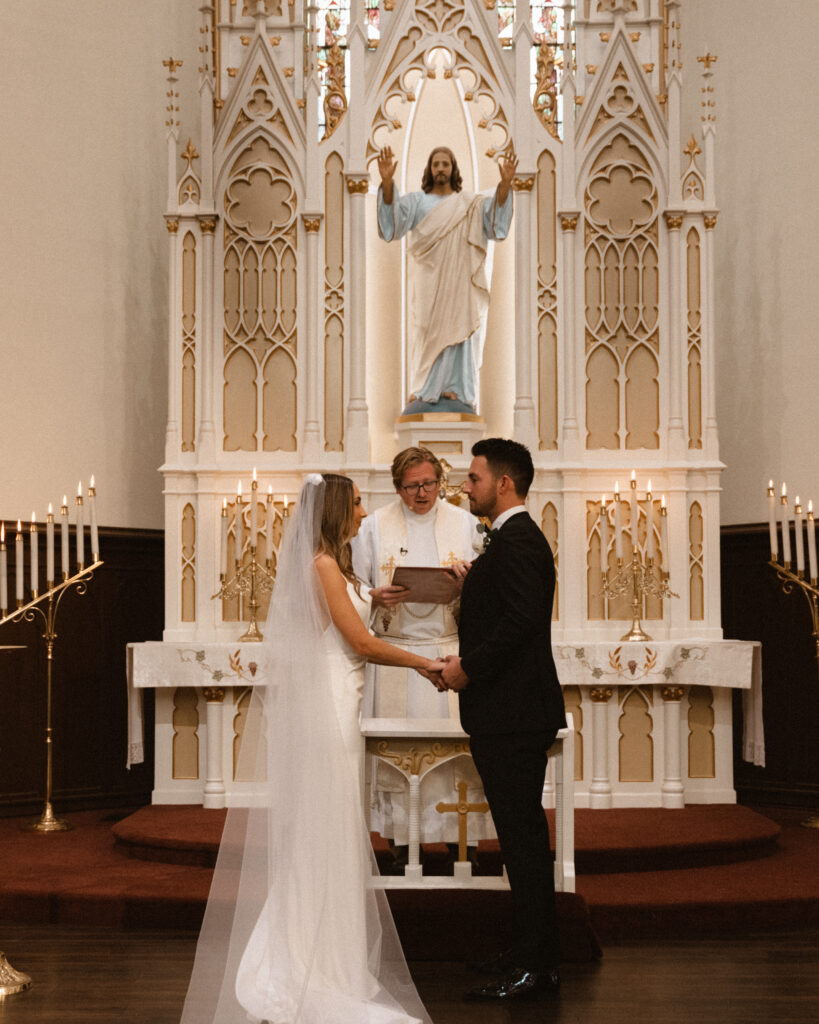 bride in cathedral veil stands with groom during church wedding ceremony
