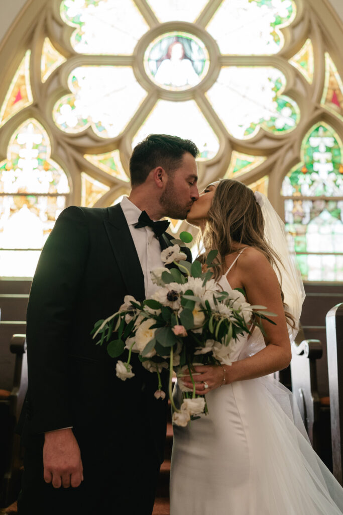 bride in modern minimalist wedding dress and groom in classic black tuxedo suit portraits in front of church stained glass windows
