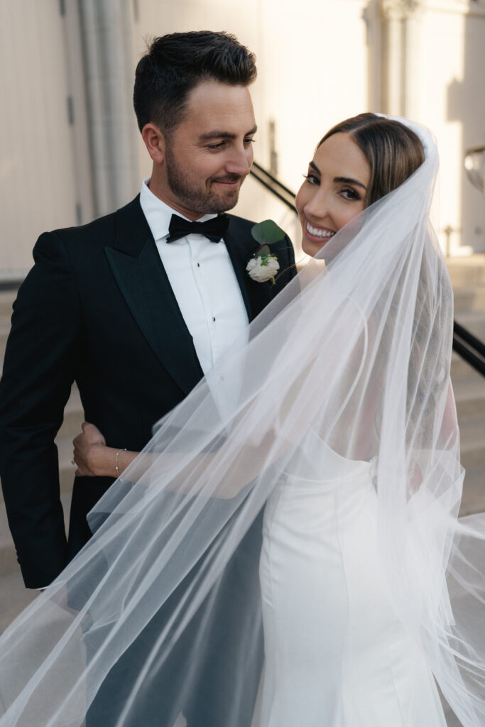 bride in modern minimalist wedding dress and cathedral veil with groom in classic black tuxedo suit take portraits on church steps