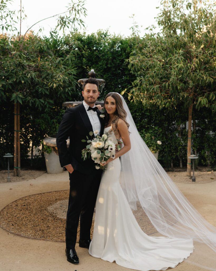 bride in modern, minimalist wedding spaghetti dress with cathedral length veil and groom in classic black tuxedo stand in front of fountain after their elegant and understated wedding in Orange County, CA