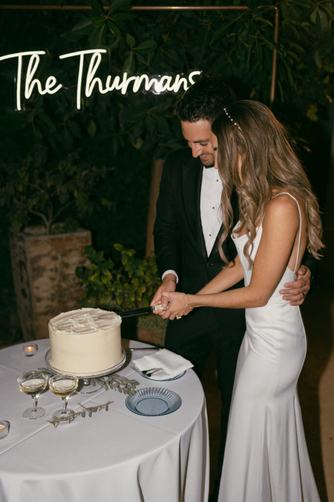 bride in modern minimalist wedding dress and groom in classic black tuxedo suit cut one tier cake with frosted "just married"