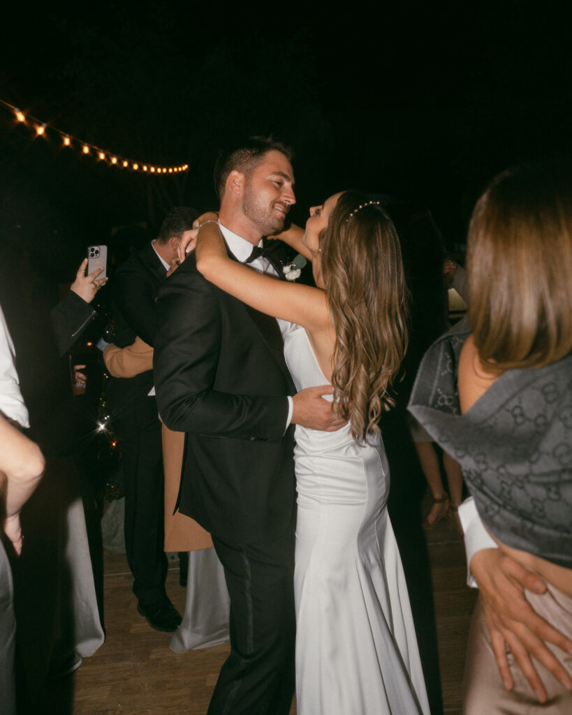 bride in modern minimalist wedding dress and groom in classic black tuxedo suit dancing with guests