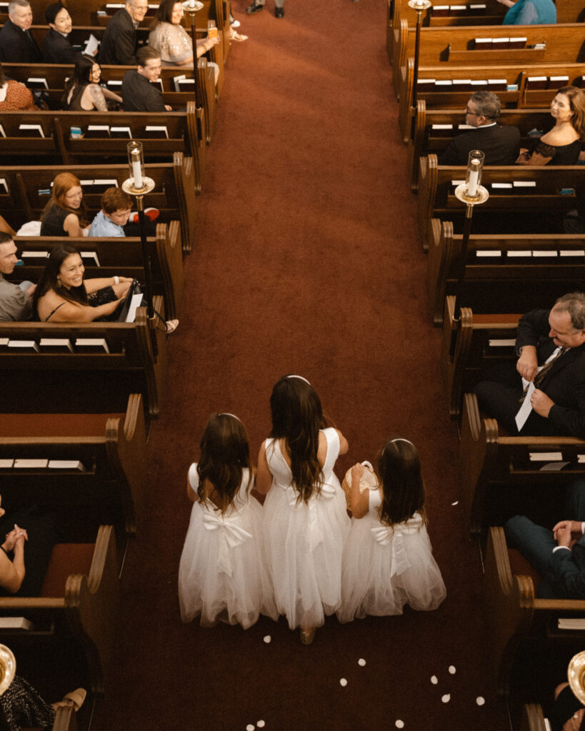 flower girls in white dresses walking down ceremony aisle during church wedding 