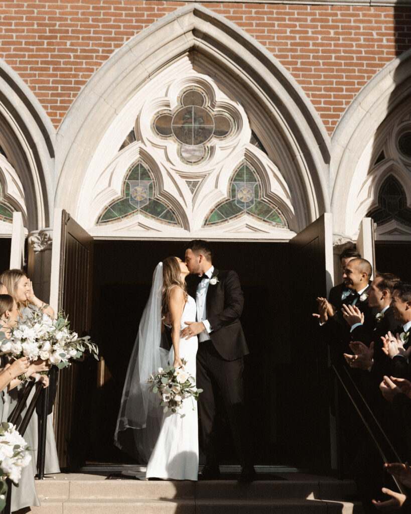  bride in modern minimalist wedding dress and groom in classic black tuxedo suit kiss outside of church steps