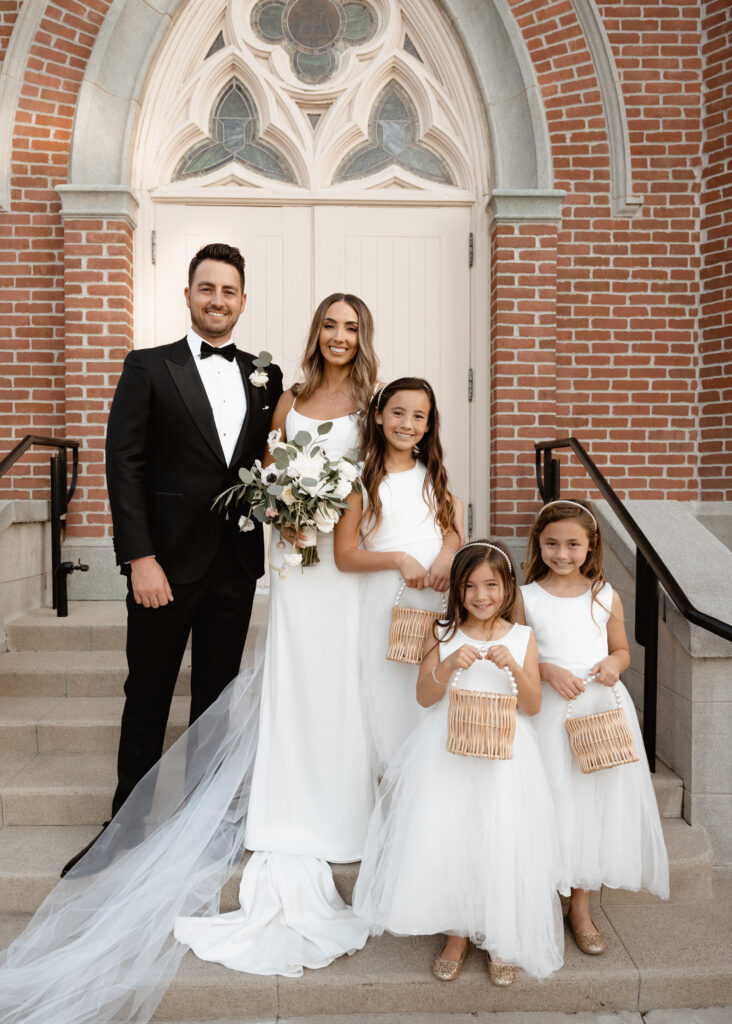 bride in modern minimalist wedding dress and cathedral veil with groom in classic black tuxedo suit take portraits on church steps with flower girls in white dresses