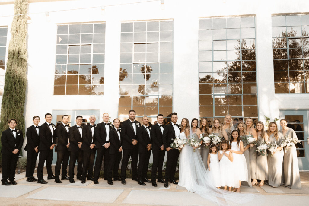 groom in classic black tuxedo suit with bowtie and bride in minimalist modern wedding dress stands with wedding party