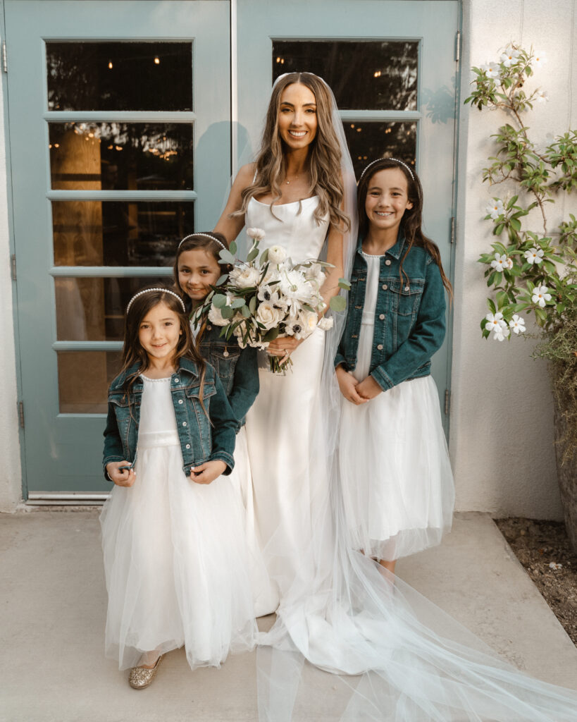 bride in modern minimalist wedding dress stands with ring girls in white dresses and jean jacket