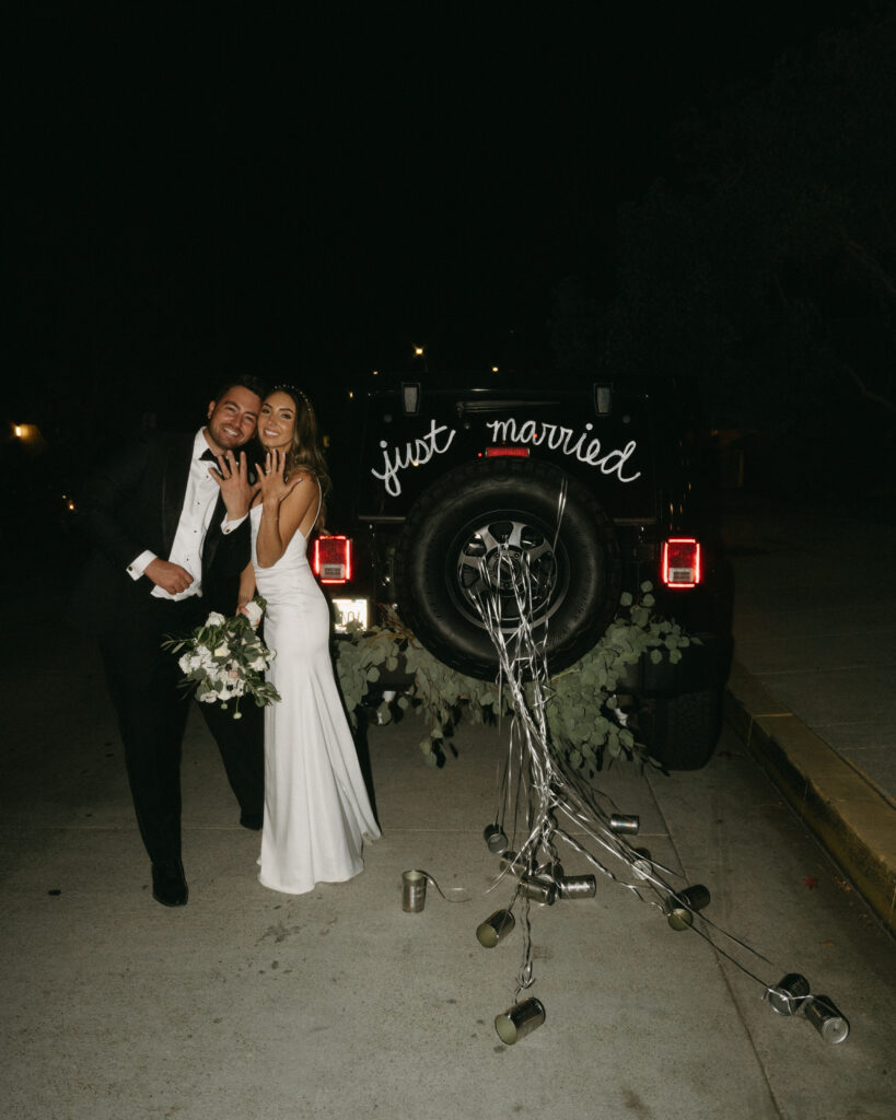 bride in modern minimalist wedding dress and groom in classic black tuxedo suit grand exit in jeep with cans and letters saying just married"