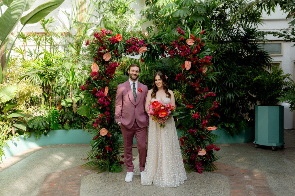 Couple stands in front of tropical inspired ceremony arch at Valentine DTLA