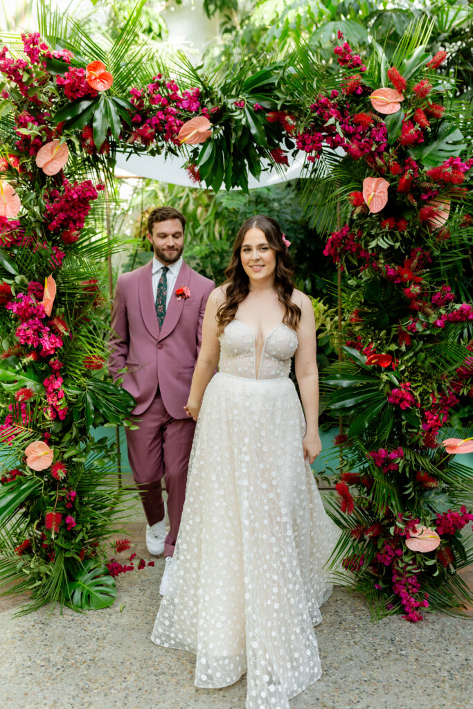 bride in strapless wedding dress with sweetheart neckline and groom in merlot colored suit with floral tie take portrait photos with tropical inspired ceremony arch