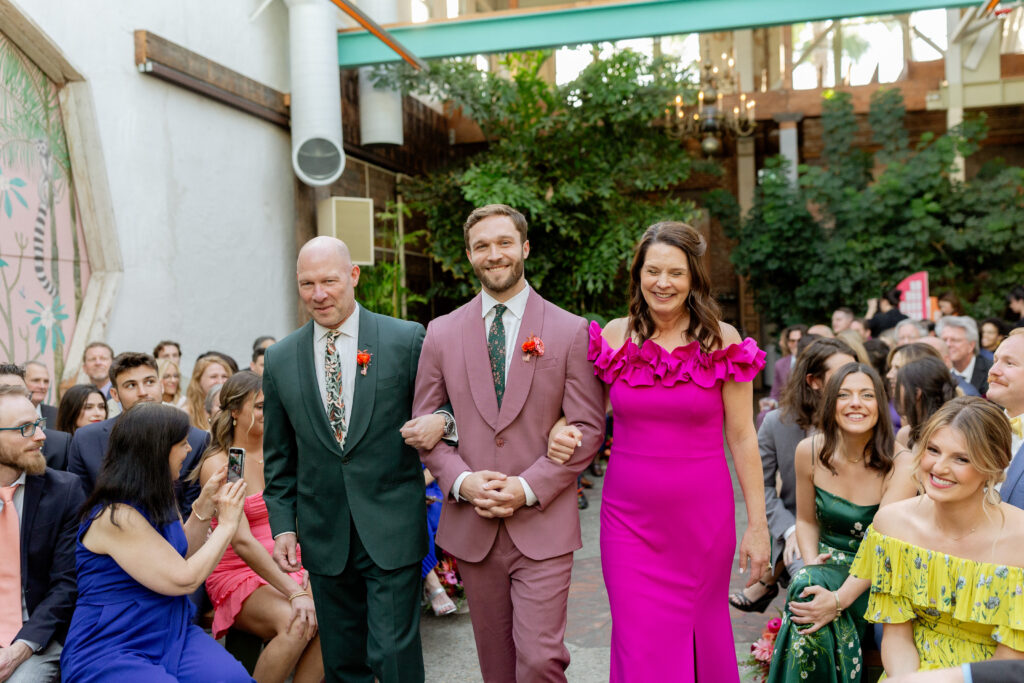 groom in merlot colored suit with floral tie walks with parents down aisle