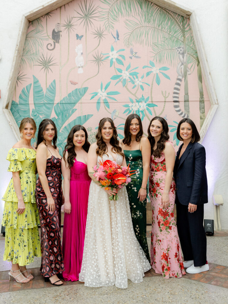bride in strapless wedding dress with sweetheart neckline and tropical floral bouquet stands with wedding party in mixed outfits