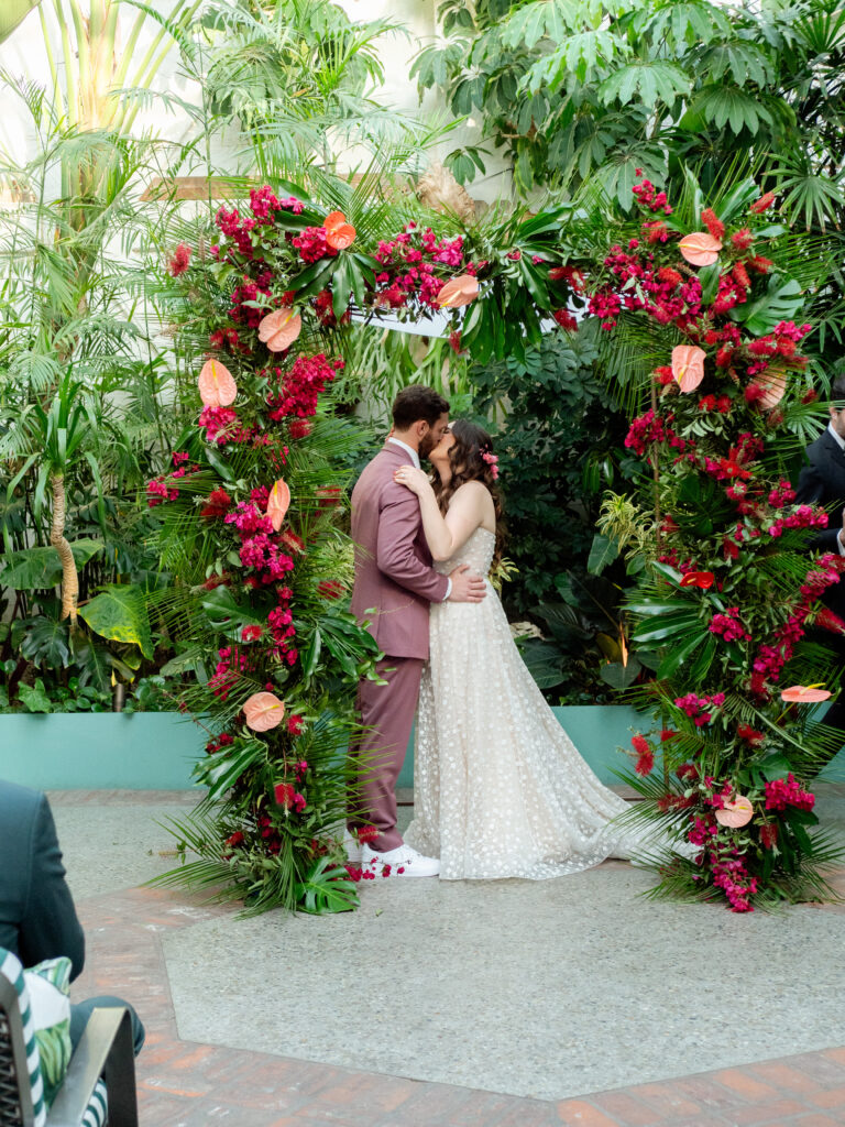 bride and groom first kiss under tropical inspired wedding ceremony arch
