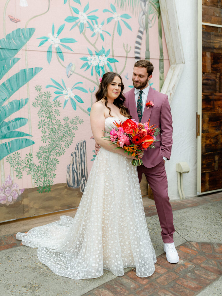 groom in merlot colored suit with white shoes and floral tie stands with bride in strapless wedding dress with sweetheart neckline and tropical floral bouquet