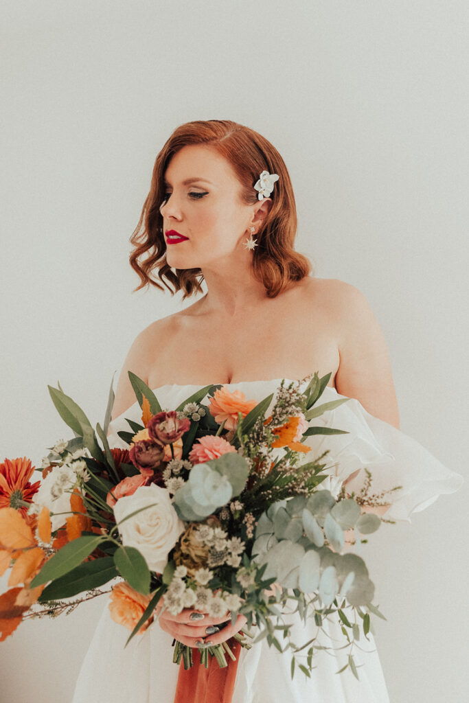 bride in modern asymmetrical off shoulder wedding dress and classic hollywood bridal look with soft waves and red lip holding colorful winter citrus inspired bridal bouquet