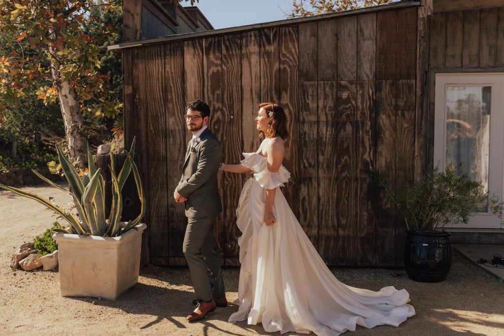 bride in modern off shoulder ruffled wedding dress with a high slit has first look with groom in warm grey suit and glasses 