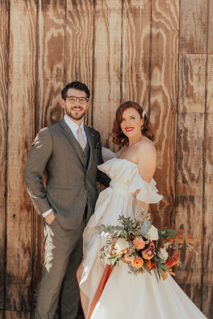 bride in modern off shoulder ruffled wedding dress with a high slit and groom with glasses and warm grey suit during portrait shots