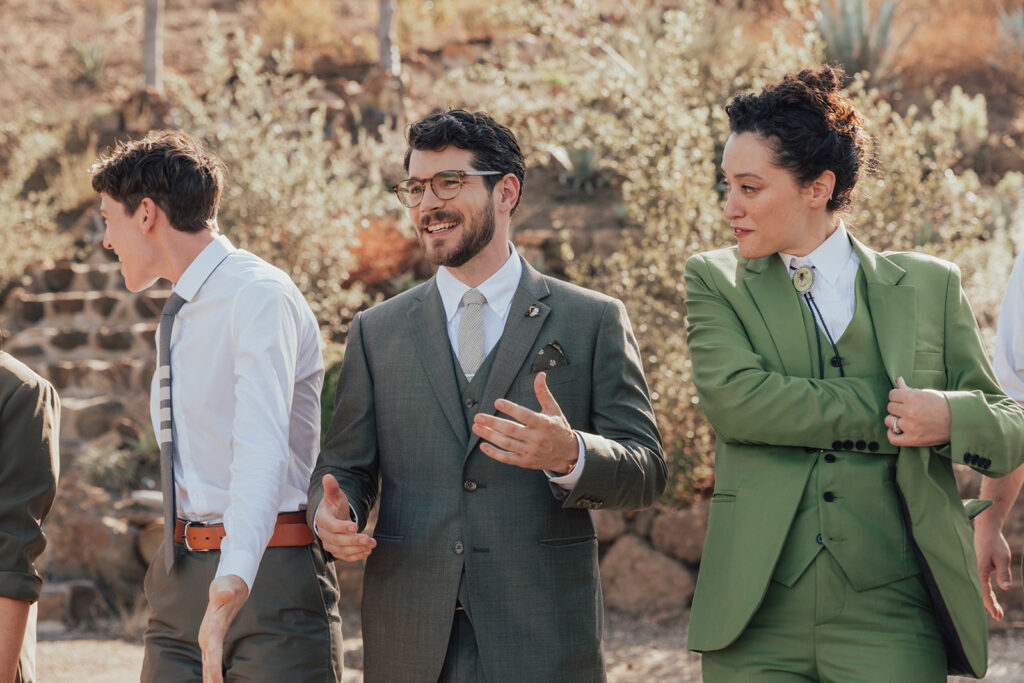 groom in warm grey suit stands with co-ed wedding party in mixed green outfits
