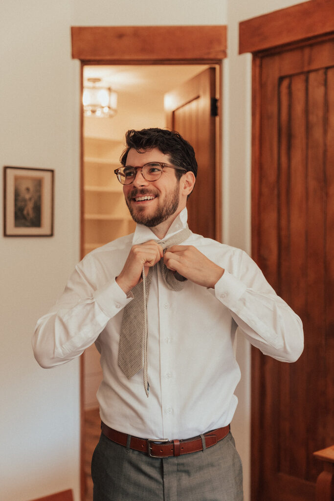 groom wearing glasses gets ready for wedding in warm grey suit with white button up and tie 