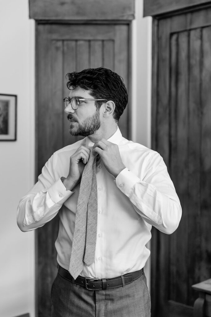 groom wearing glasses gets ready for wedding in warm grey suit with white button up and tie 