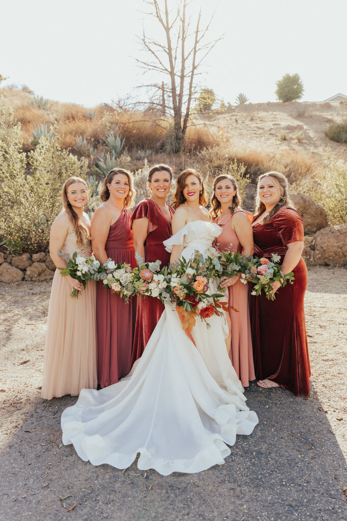 bride in modern off-shoulder ruffled wedding dress with high slit stands with wedding party wearing mixed red dresses