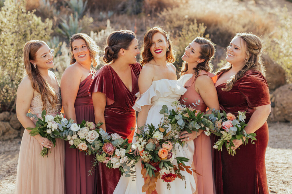 bride in modern off-shoulder ruffled wedding dress with high slit stands with wedding party wearing mixed red dresses