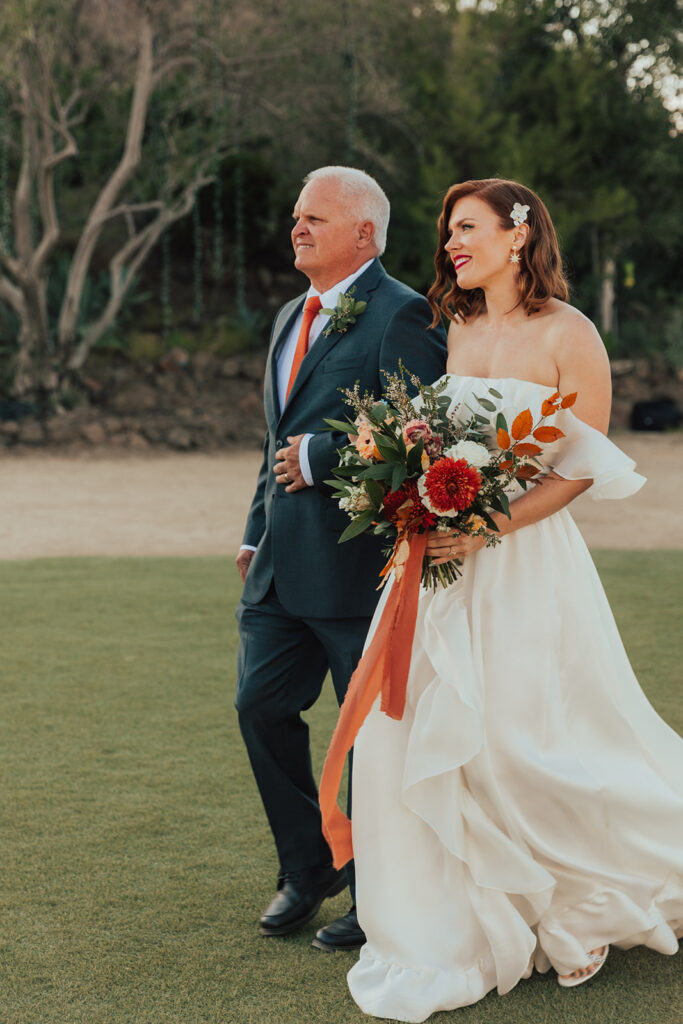 bride in modern ruffled off-shoulder wedding dress with high slit holds colorful bouquet while walking down ceremony aisle