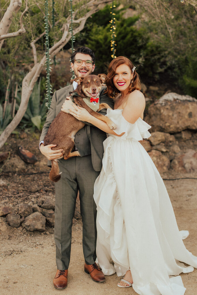 bride in modern asymmetrical off-shoulder wedding dress with groom in warm grey suit and glasses takes photo with dog in bow tie during citrus inspired wedding