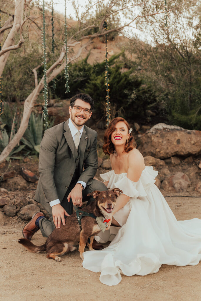 bride in modern ruffled off-shoulder wedding dress with high slit and groom with glasses wearing a warm grey suit with tie take portrait shots with dog