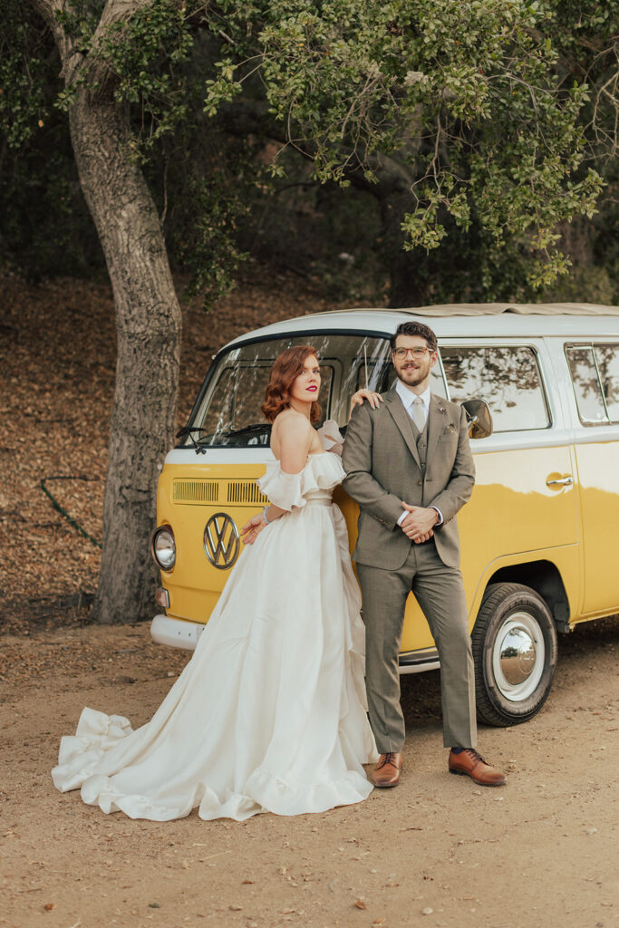 bride in modern ruffled off-shoulder wedding dress with high slit and groom with glasses wearing a warm grey suit with tie take portrait shots with colorful vintage Volkswagens
