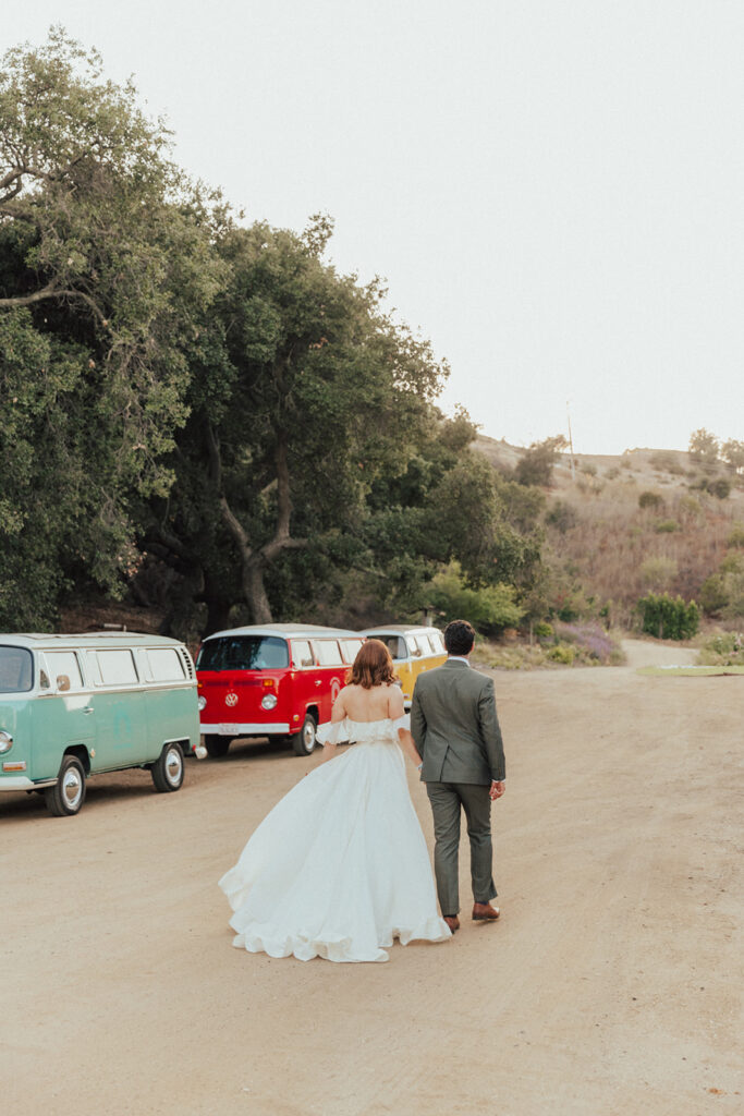 bride in modern ruffled off-shoulder wedding dress with high slit and groom with glasses wearing a warm grey suit with tie take portrait shots with colorful vintage Volkswagens