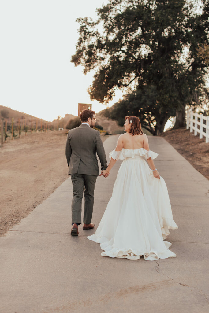 bride in modern ruffled off-shoulder wedding dress with high slit and groom with glasses wearing a warm grey suit with tie take portrait shots during sunset