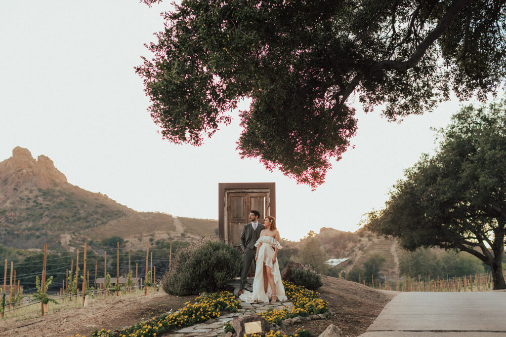 bride in modern ruffled off-shoulder wedding dress with high slit and groom with glasses wearing a warm grey suit with tie take portrait shots during sunset