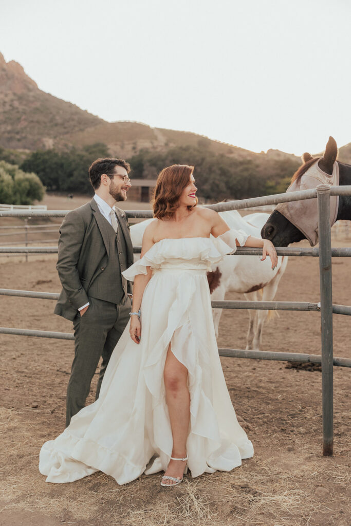 bride in modern ruffled off-shoulder wedding dress with high slit and groom with glasses wearing a warm grey suit with tie take portrait shots with horses during sunset