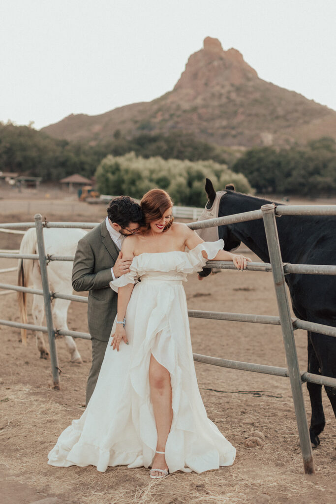 bride in modern ruffled off-shoulder wedding dress with high slit and groom with glasses wearing a warm grey suit with tie take portrait shots with horses during sunset