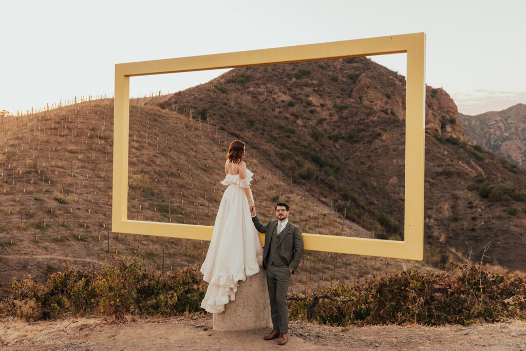bride in modern ruffled off-shoulder wedding dress with high slit and groom with glasses wearing a warm grey suit with tie take portrait shots with yellow frame during sunset