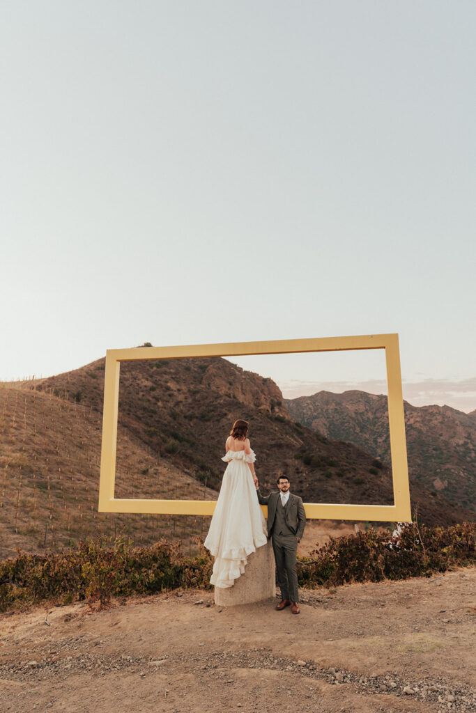 bride in modern ruffled off-shoulder wedding dress with high slit and groom with glasses wearing a warm grey suit with tie take portrait shots with yellow frame during sunset