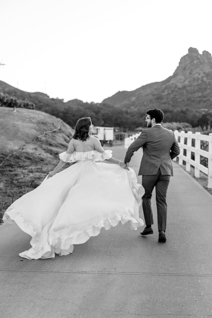 bride in modern ruffled off-shoulder wedding dress with high slit and groom with glasses wearing a warm grey suit with tie take portrait shots  during sunset