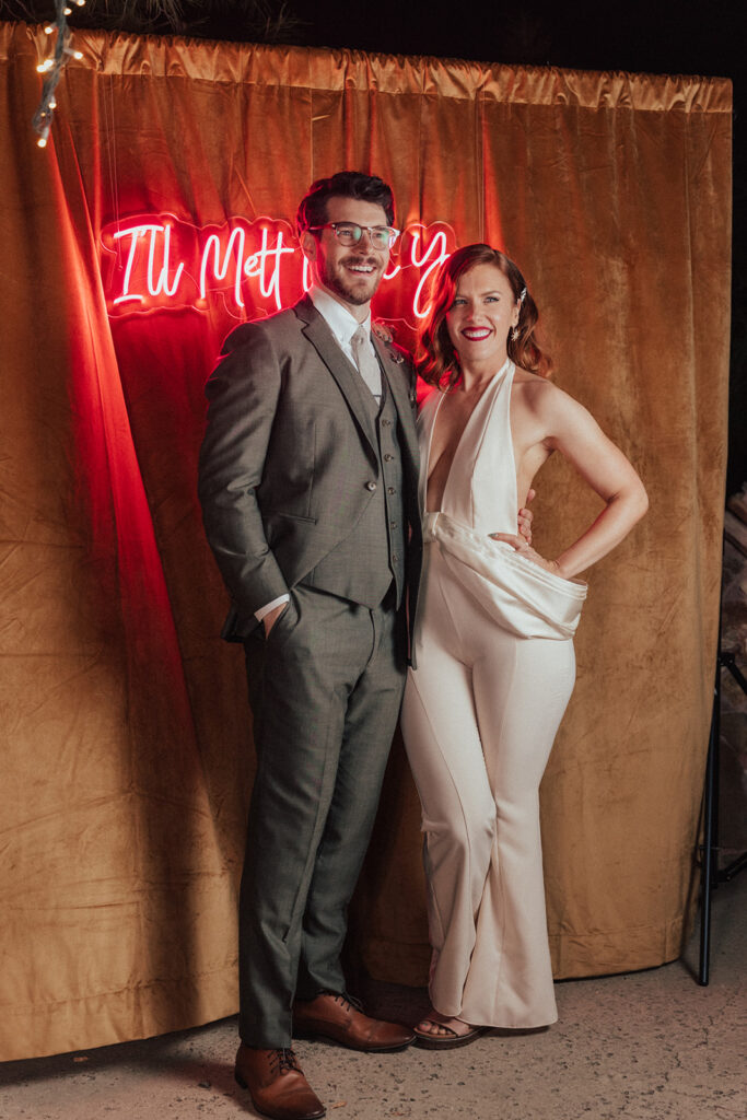 bride in modern wedding reception jumpsuit with groom take photos with neon letter back drop
