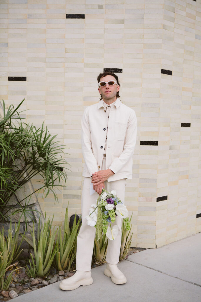 groom in all white wedding outfit and sunglasses holding purple, green and white bouquet