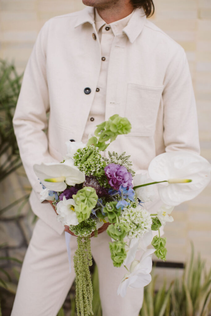 groom in all white wedding outfit and sunglasses holding purple, green and white bouquet