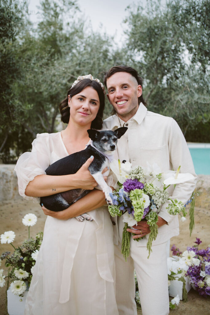 bride in modern wedding dress with sheer overlay and satin headband stands with groom in white suit holding a purple and green bouquet and their dog during their laidback, cool wedding at the Ace Hotel 