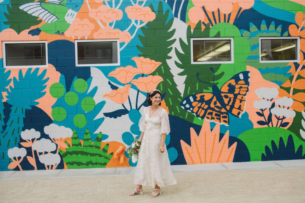 bride in modern wedding dress with sheer overlay and satin headband holds purple, green and white bouquet in front of colorful wall mural at Ace Hotel 