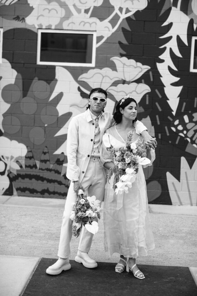 first look with bride in modern wedding dress with sheer overlay and satin headband and groom in all white outfit with both holding bouquets in front of colorful wall mural 