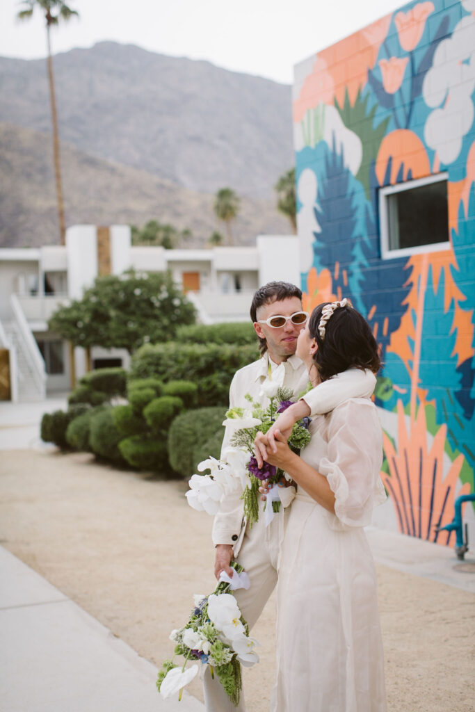 first look with bride in modern wedding dress with sheer overlay and satin headband and groom in all white outfit with both holding bouquets in front of colorful wall mural 