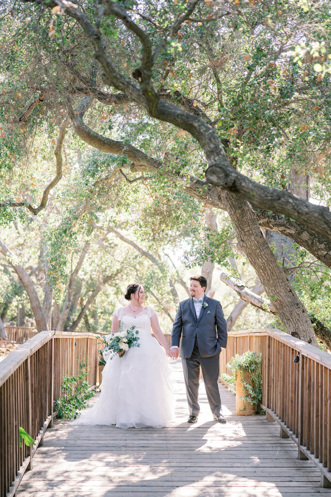 bride in organza ballroom wedding dress and groom in dark grey suit and sage bowtie hold hands at Calamigos Ranch during light and airy wedding 