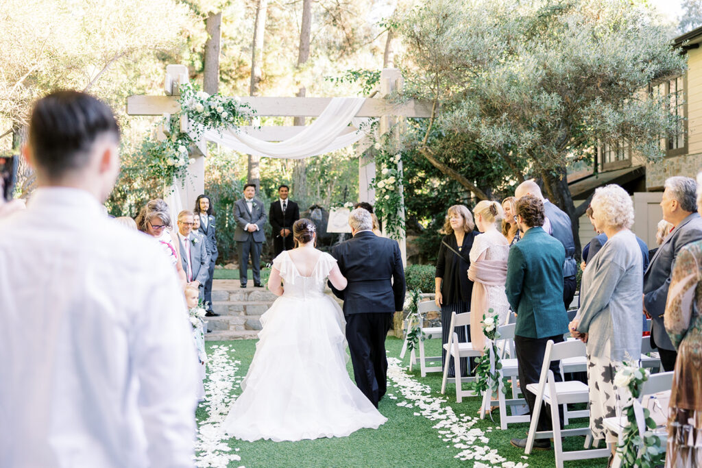 bride walking down aisle during light and airy wedding ceremony at Calamigos Redwood Room with white drapery and flowers