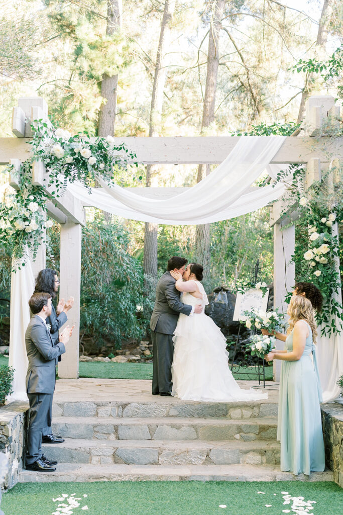 bride and groom first kiss during light and airy wedding ceremony at Calamigos Redwood Room with white drapery and flowers