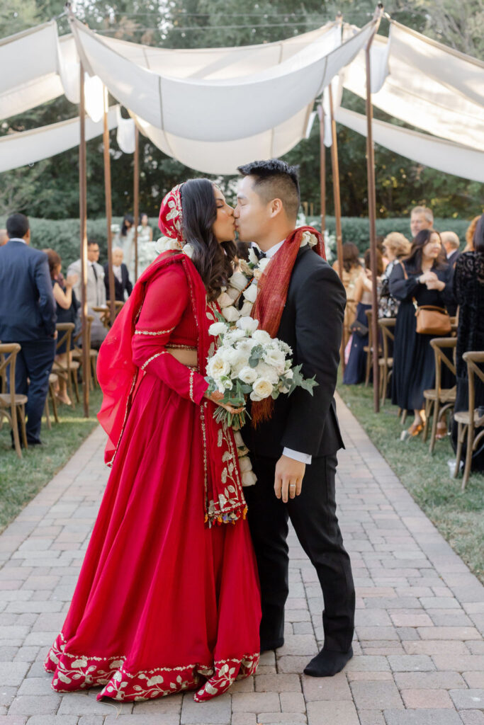 bride in red saari and groom in black tuxedo kiss during recessional 