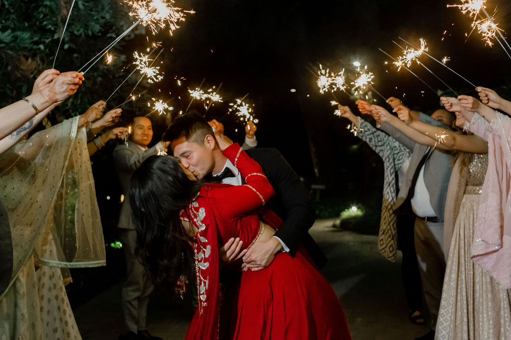 bride in red saari and groom in black tuxedo have grand exit with sparklers